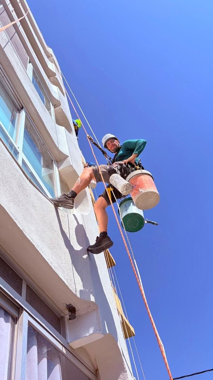 Pintor de AV Mantenimiento Integral pintando una fachada descolgado con cuerdas y cubos de pintura